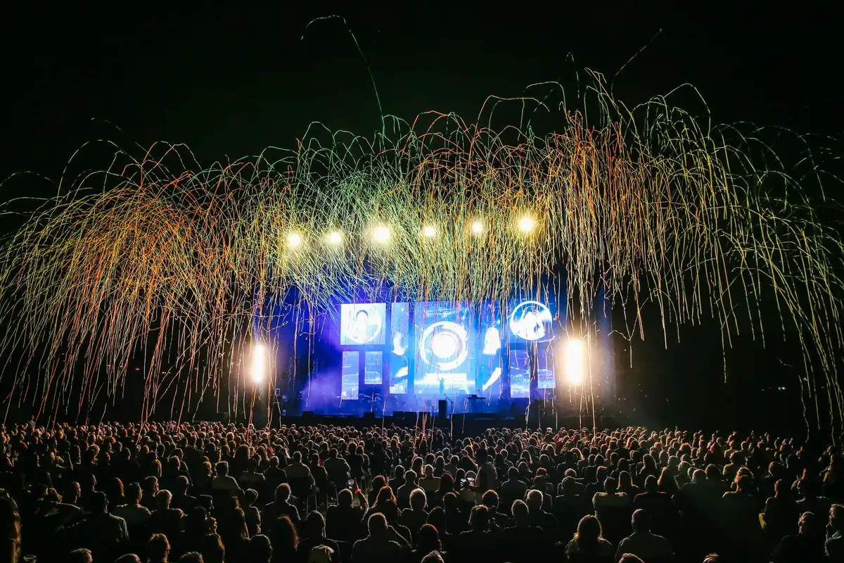 Silhouetted performer on stage with raised arms facing an engaged crowd under warm concert lights in a mid-size venue