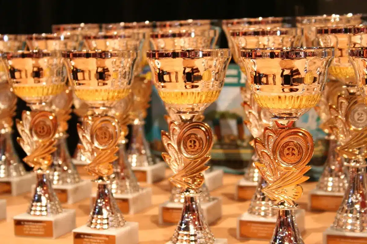 Empty awards stage under soft spotlights, a single microphone stand on center stage and a lone trophy on the floor, audience seats dark; evokes an upcoming live ceremony