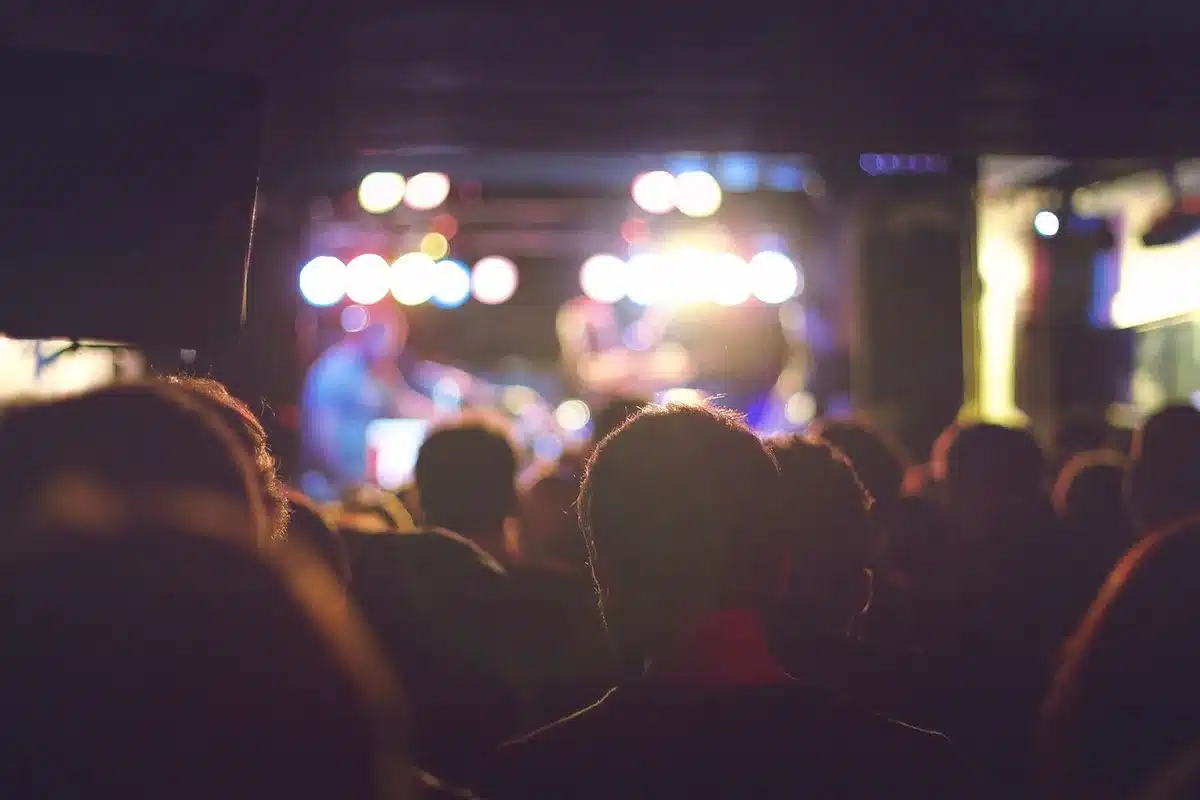 Audience silhouettes in a darkened arena, phone screens glowing as lights dim and a stage silhouette waits in the distance