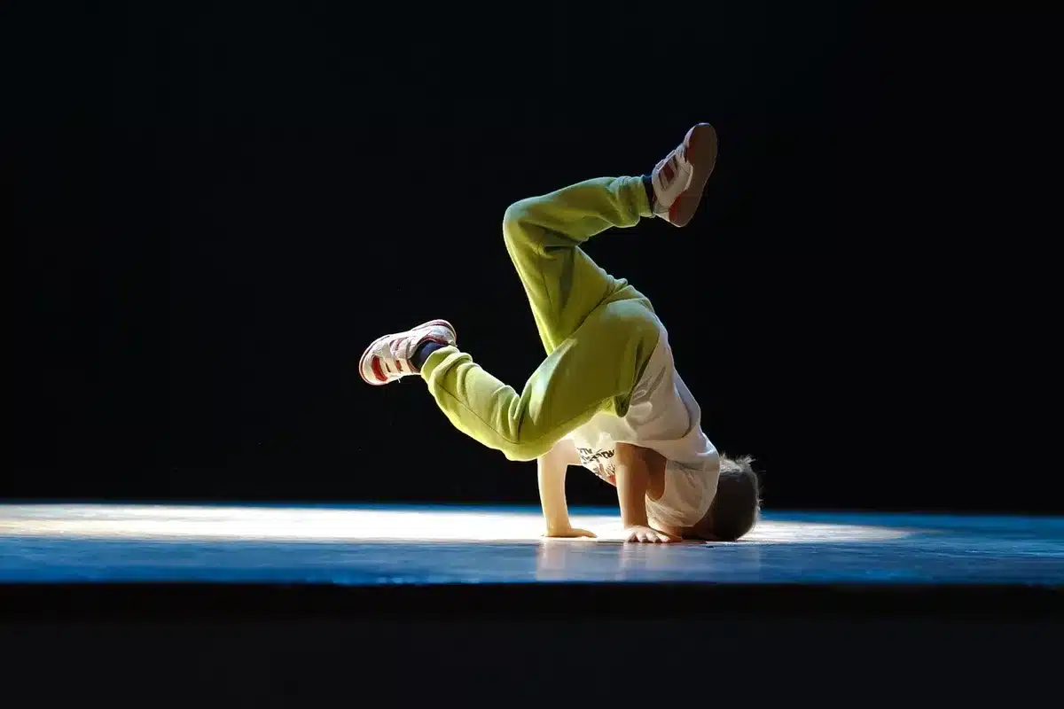 Two anonymous figures mid-rehearsal, blurred in motion under studio lights, one reaching toward the other across a gym floor — dynamic, unbranded movement