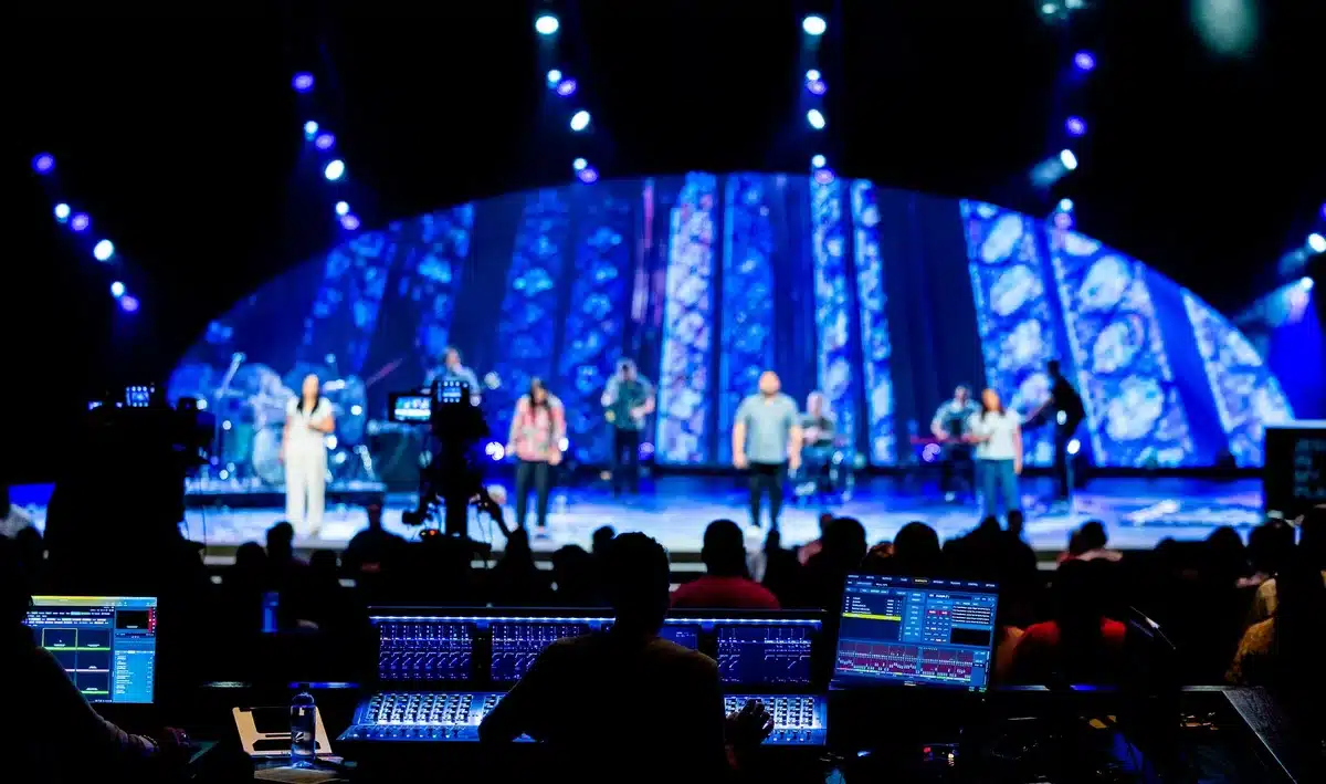 An elevated multi-tiered concert stage with a large unbranded speaker prop, silhouetted backing dancers on catwalks and a lone anonymous figure atop the platform — tension