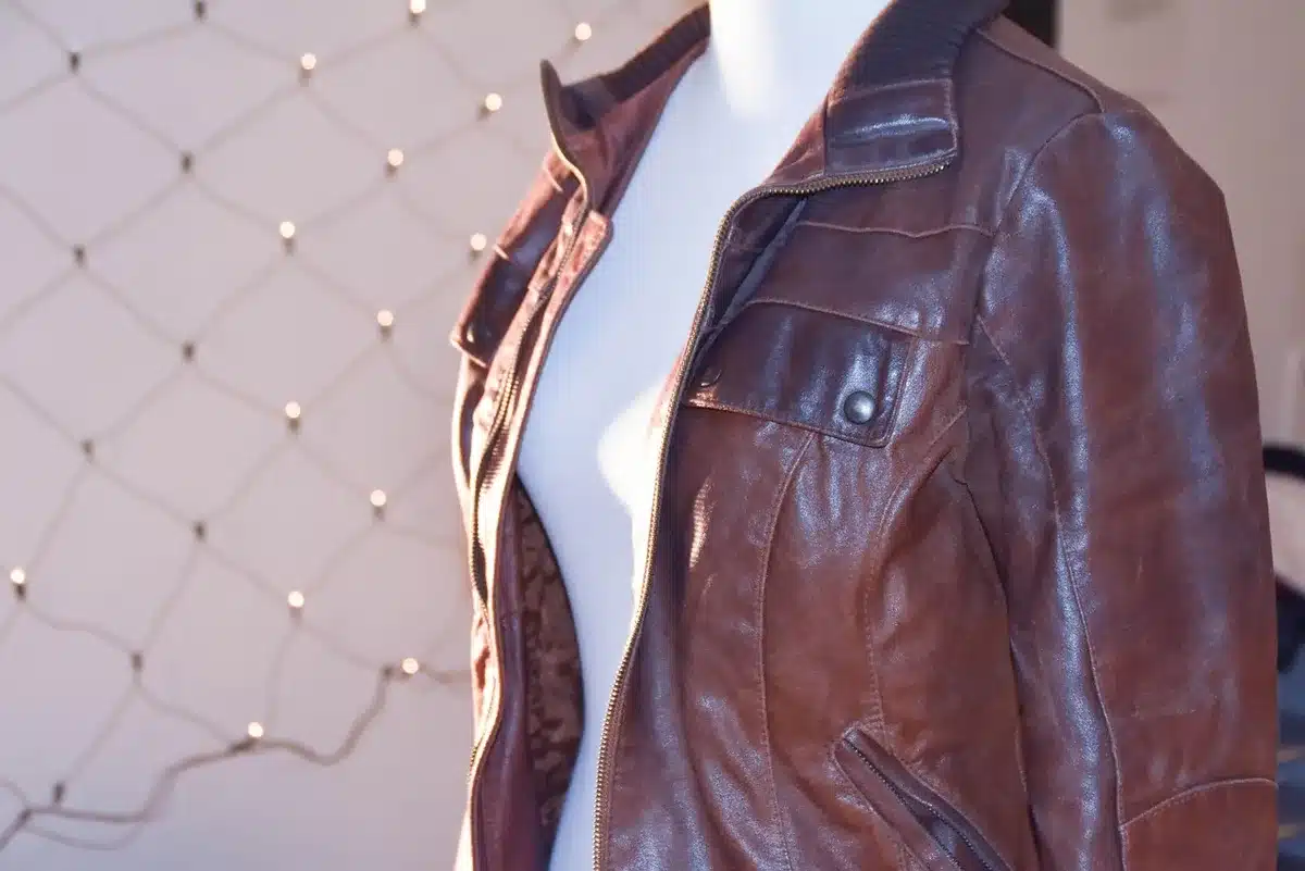A stand-collar jacket in warm suede or leather, photographed from the front, hung on a minimalist hanger against a neutral backdrop, showing the distinctive upright collar detail