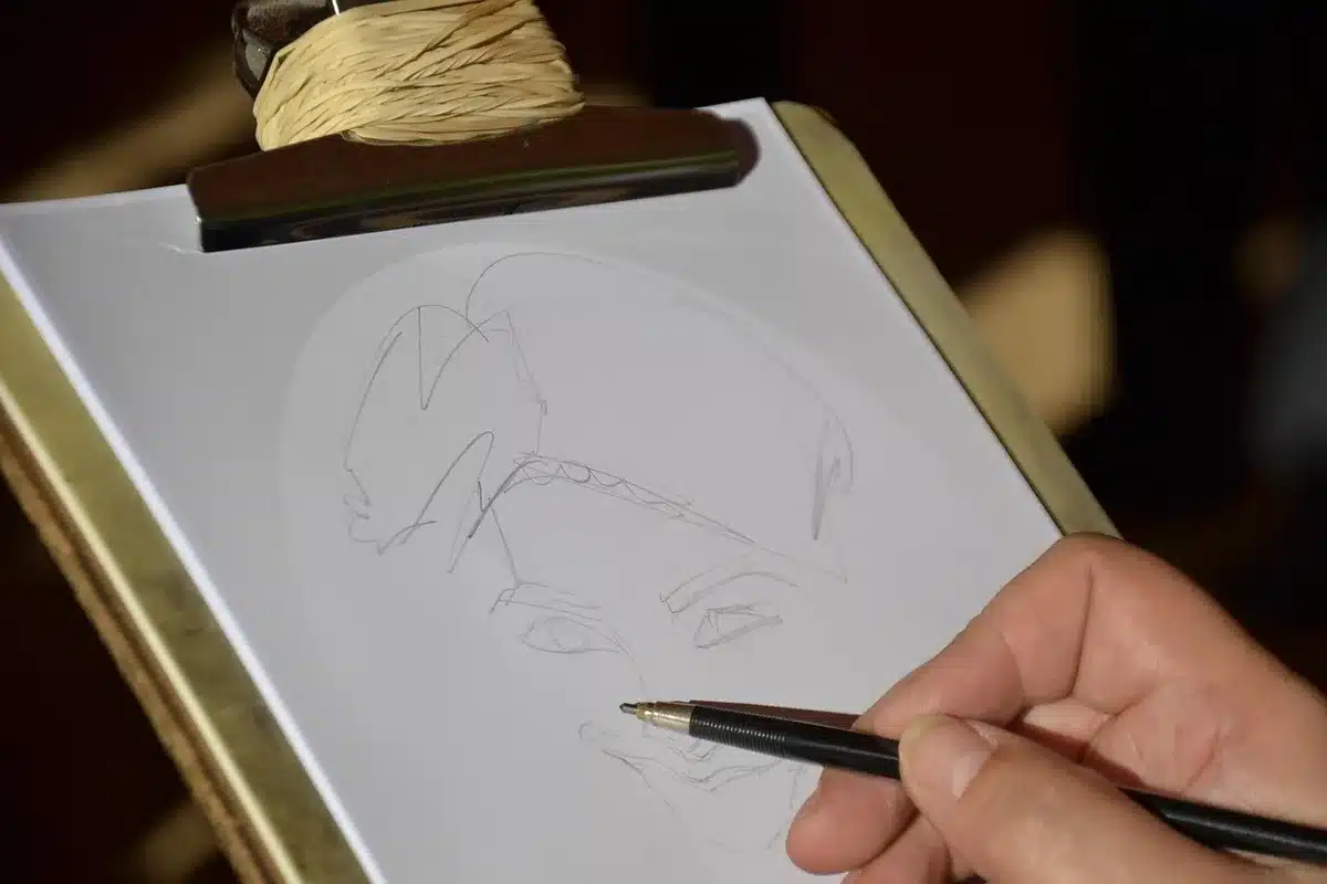 An artist's hand drawing with a pen on paper, surrounded by sketches and creative materials