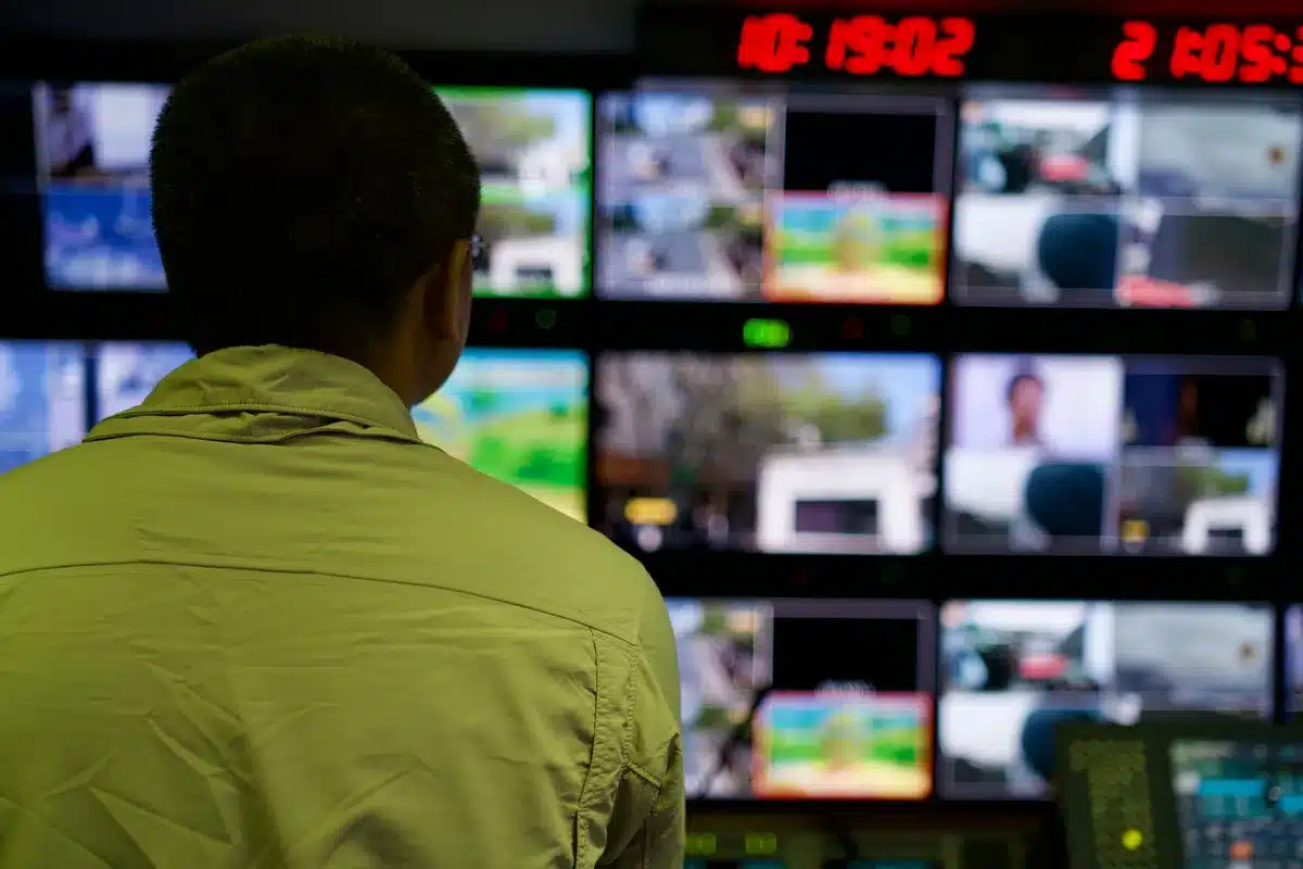 An empty television control room with stacked broadcast monitors showing generic channel id cards, a dark console in the foreground and a poster board reading "Live Event" (no logos) on the wall