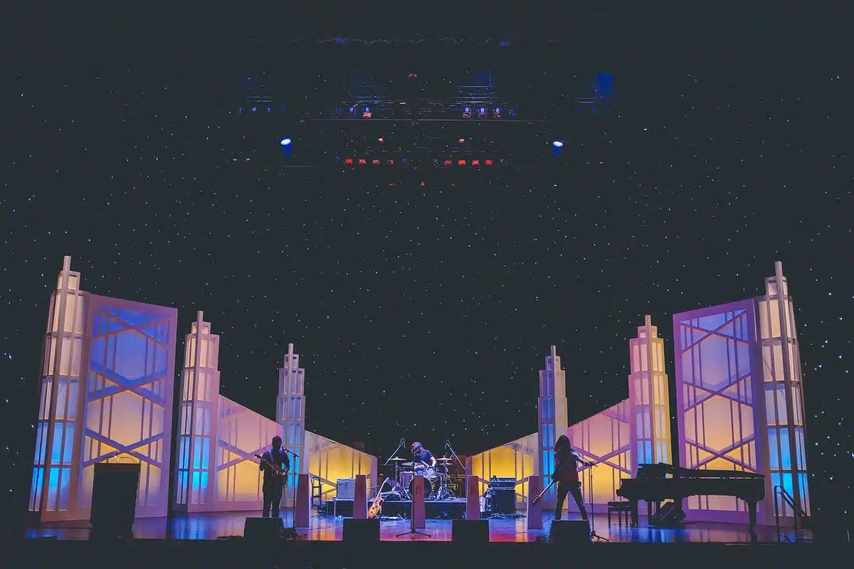 A dim stage with a single empty microphone stand under a spotlight, fog drifting low across the floor and a distant LED wall glowing — no logos, no performers