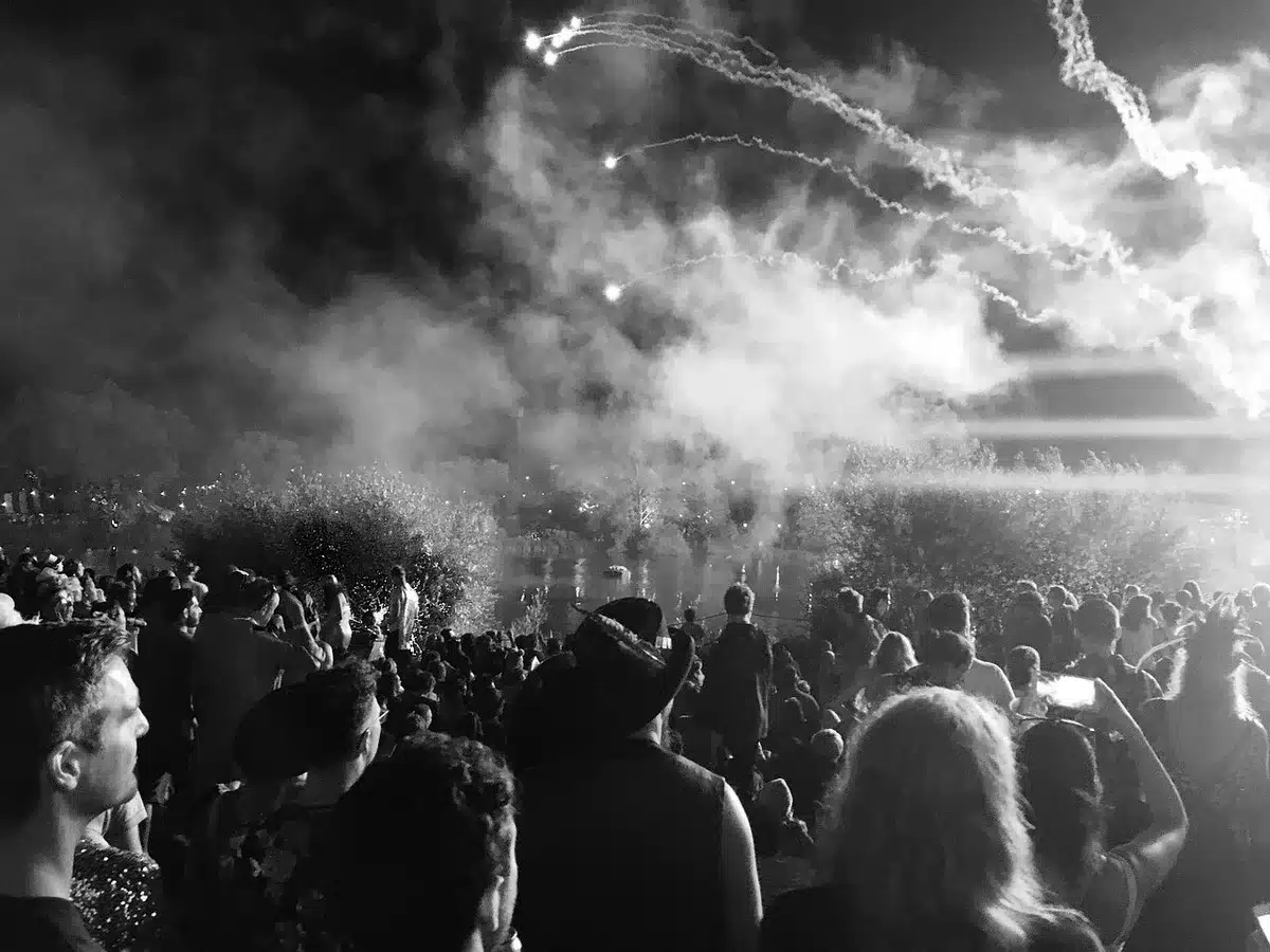A wide shot of a packed festival field under stage lights, anonymous crowd silhouettes raising hands toward a distant stage with smoke and confetti in the air