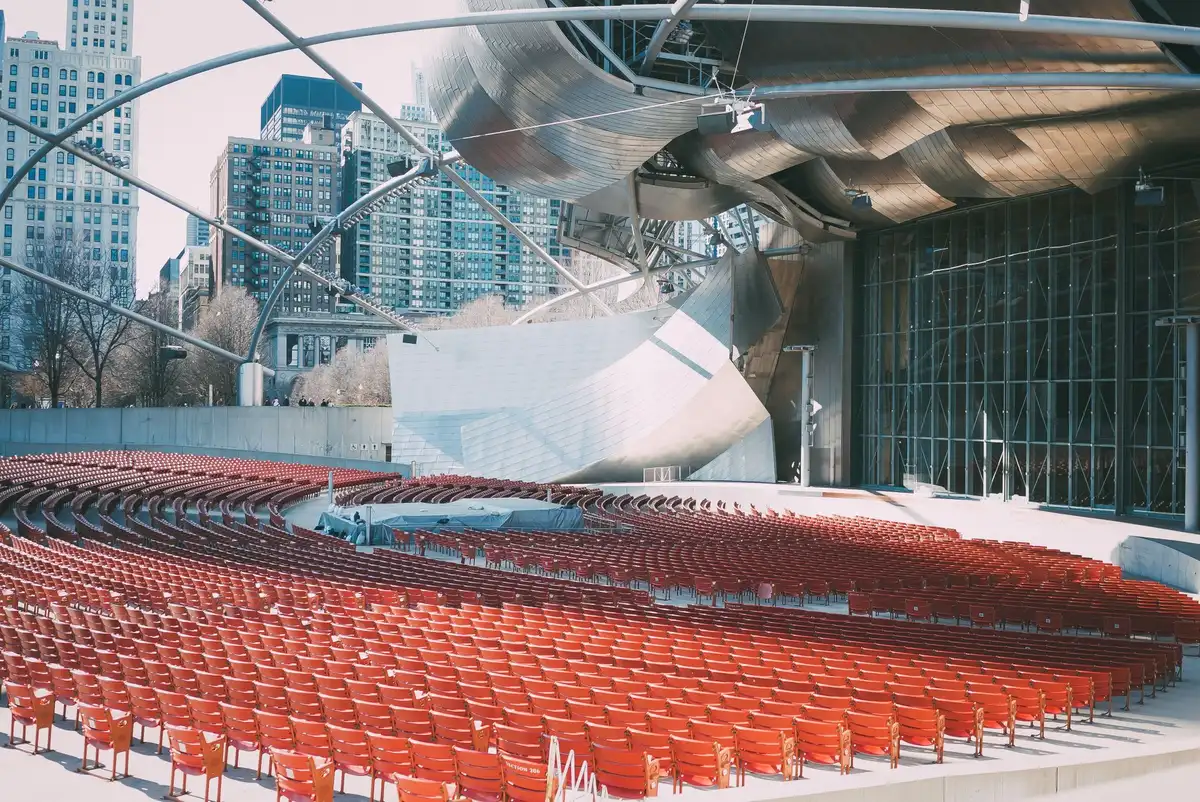 Empty outdoor amphitheater at dusk with stage lights glowing, rows of empty lawn seating and a distant city skyline