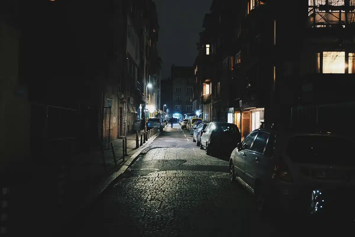 A dimly lit alley with a lone anonymous figure in a hooded jacket standing beneath a fire escape, rain-slick pavement reflecting neon lights, atmosphere of tension and pursuit