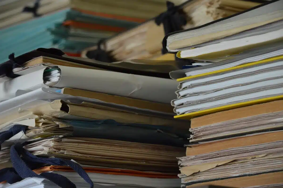A close-up of an anonymous desk scattered with case files, a worn notepad, a black pen and a low-lit evidence photo, suggesting a detective’s investigation