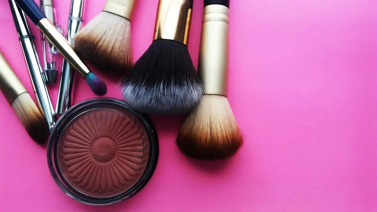 anonymous backstage table with brushes, tubes of gloss, loose pigment pots and a stray hair clip, hands reaching in to finish a look