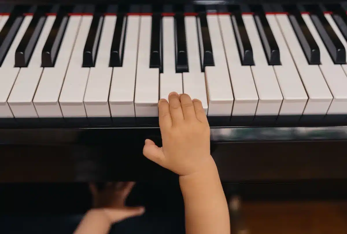 An empty piano bench facing a child-sized keyboard under a spotlight, a lone pair of small hands resting on the keys, anonymous and out of frame