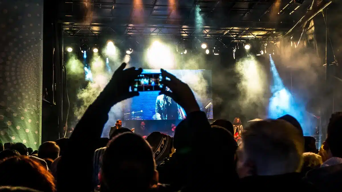 A crowded music venue with silhouettes of anonymous patrons facing a lit stage, raised hands and movement, warm stage lights and cool ambient lighting creating contrast