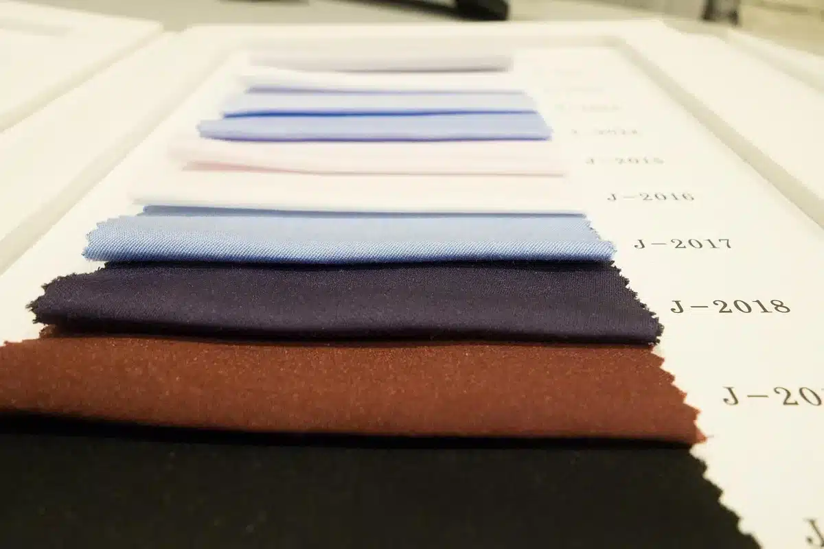 A young designer's hands working with fabric samples and origami-folded garments, natural light streaming across a wooden table with sketches and color swatches.