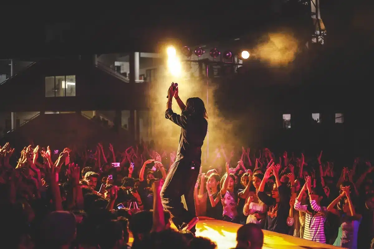 A wide, anonymous crowd under string lights at an outdoor festival, hands raised toward a distant illuminated stage; dusk sky and stage glow — a festival atmosphere, awe
