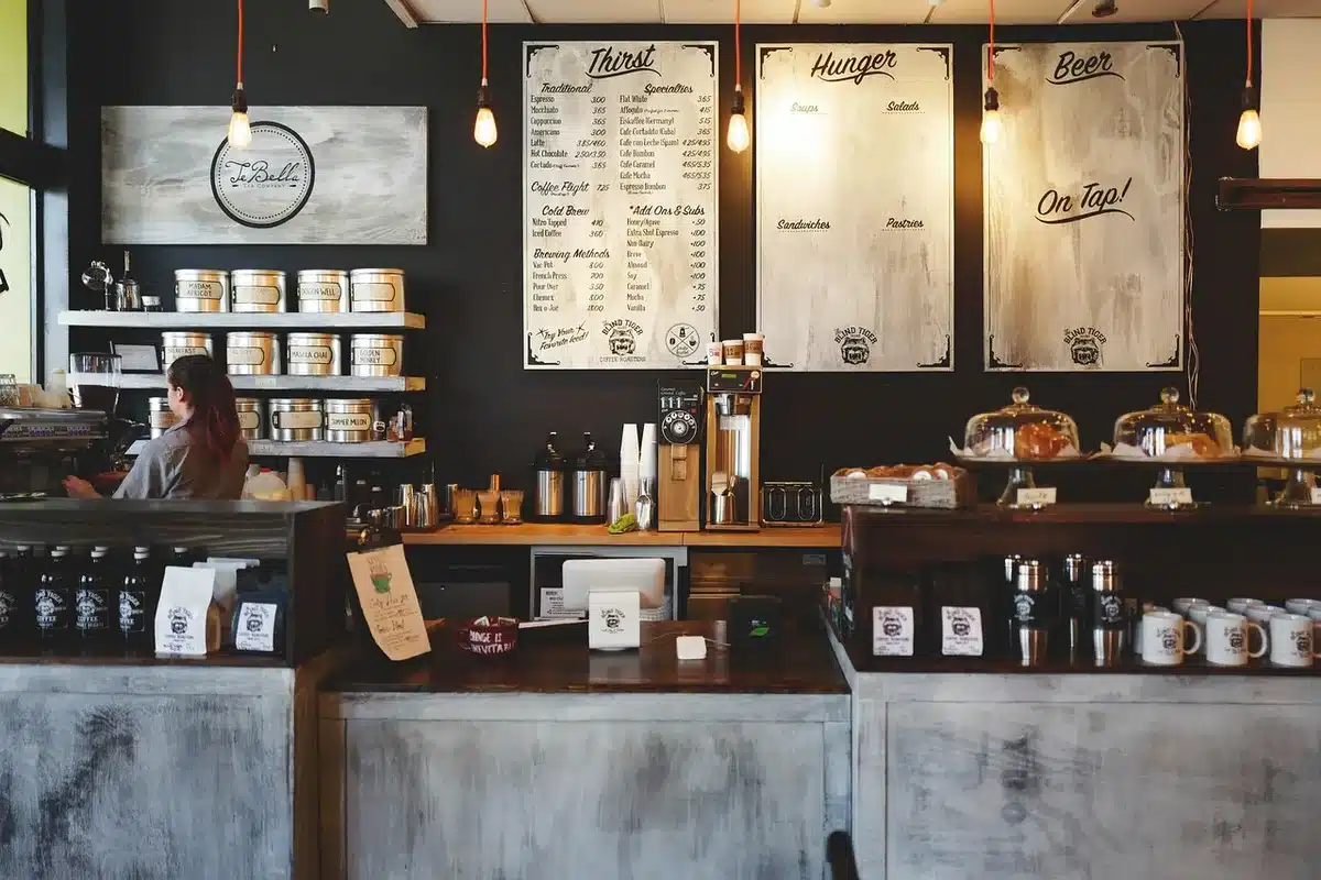 A quiet independent comic shop interior with rows of unbranded graphic novels on wooden shelves, a handwritten event flyer board in the background, and a single checkout counter — evokes local shop community life