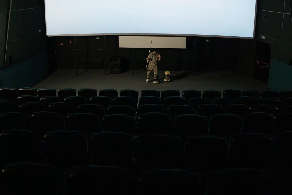 A dimly lit empty screening room with one solitary projector beam cutting across empty seats, a cancelled screening notice on a nearby counter, hinting at funding uncertainty