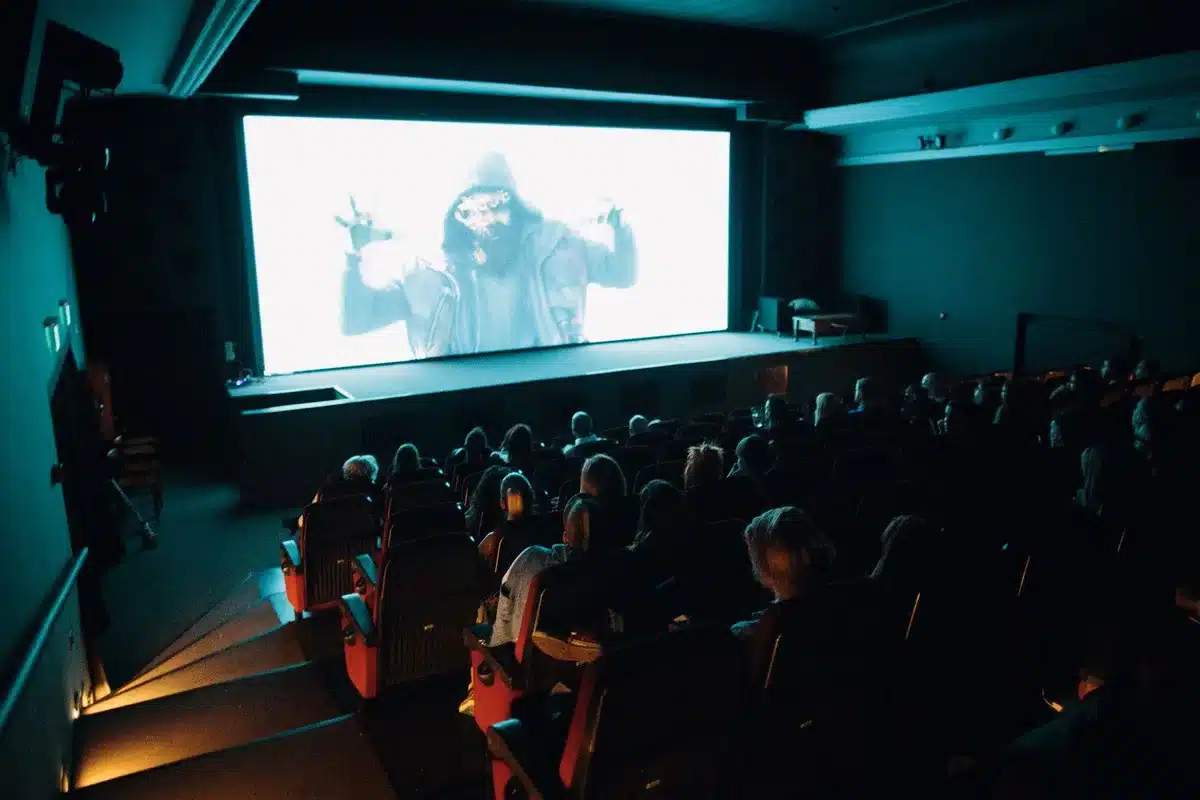 A dim, anonymous crowd of viewers silhouetted against a cinema screen, some heads turned away mid‑reaction — an atmosphere of shock and fascination