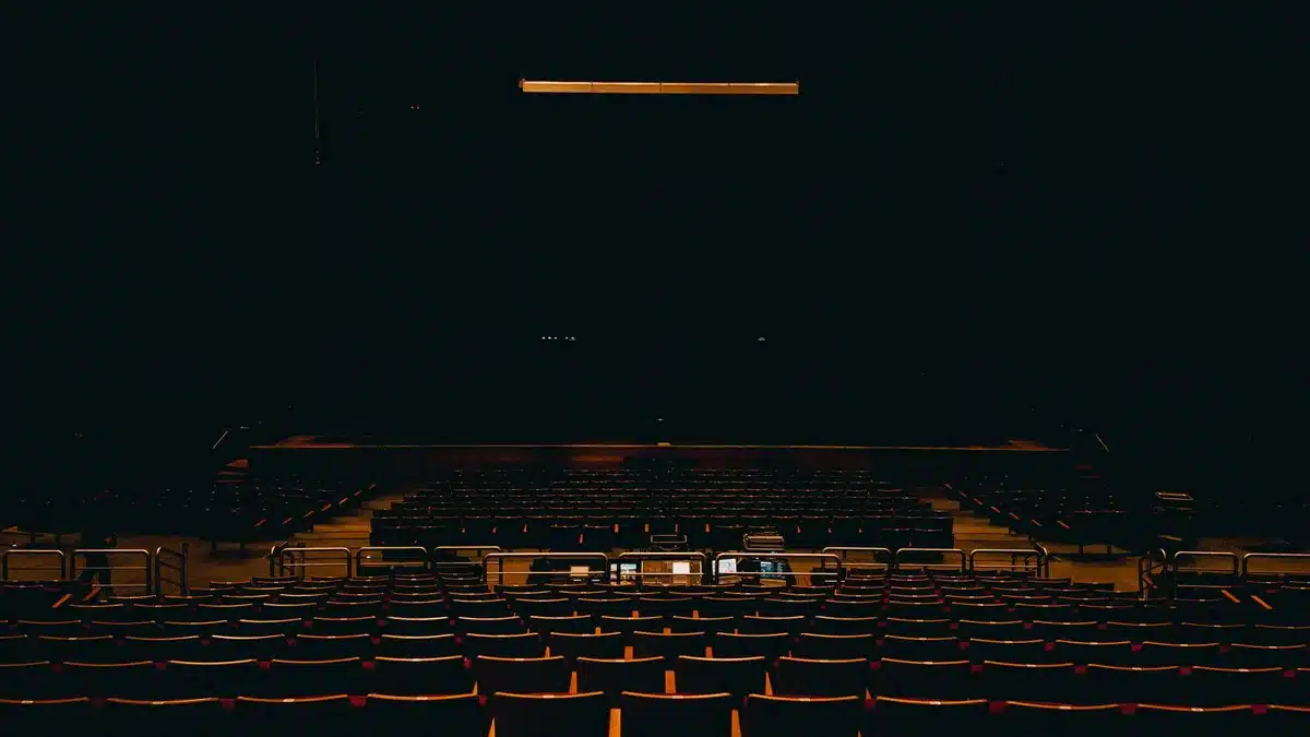 Empty theater stage with a single spotlight on an unbranded microphone stand and coiled cable, dust motes visible in the beam