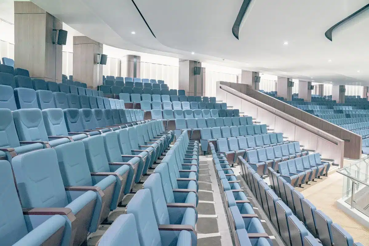 Empty rows of an auditorium facing a lit stage and a lone chair beneath a spotlight, no people visible