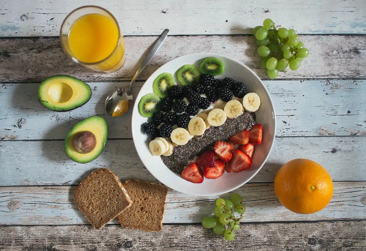 A rustic wooden kitchen table scattered with a variety of anonymous unbranded fruit and vegetables, one peeled banana half-hidden under a newspaper, knife and a single empty cereal bowl nearby