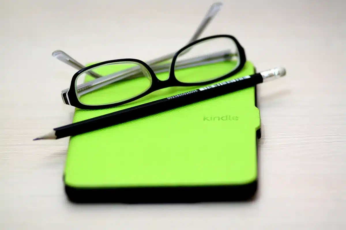 A close-up of an open e-reader resting on a coffee table beside a pair of anonymous earbuds, a paperback and a notepad visible out of focus