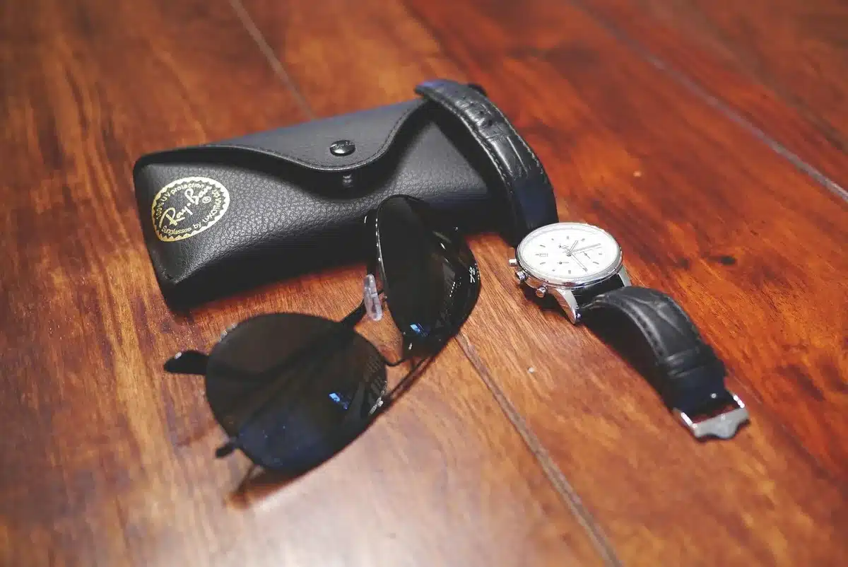 A flatlay of anonymous personal accessories — a simple unbranded pendant on a dish, a plain phone case, and an insulated tumbler — arranged on a wooden table