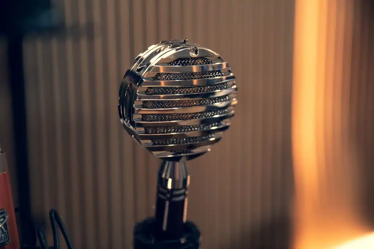 A dimly lit empty stage with a single vintage microphone on a stand under one hard spotlight, dust motes visible in the beam, an anonymous shadowed chair behind it